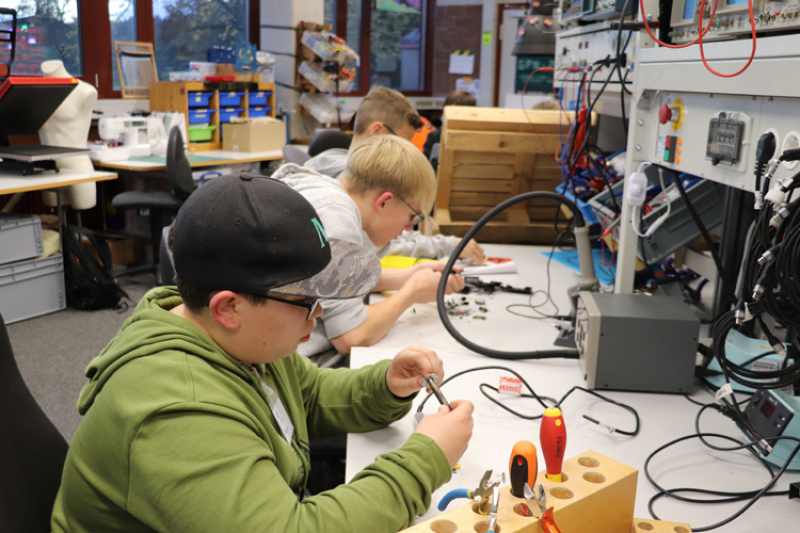  In einem Werkraum sitzen mehrere Jungen an Tischen und arbeiten an kleinen Elektronik- oder Bastelprojekten.