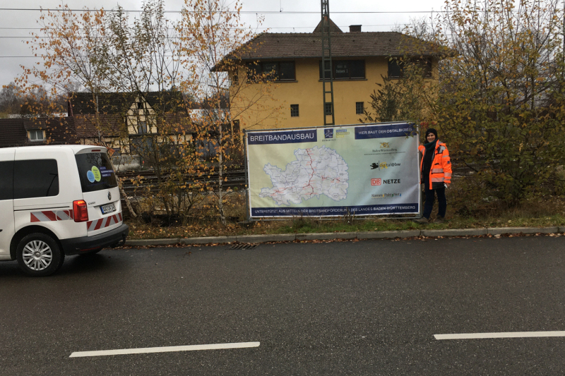 Ein Mitarbeiter der Vermessung steht am Straßenrand neben einem großen Schild zum Breitbandausbau Ostalbkreis.