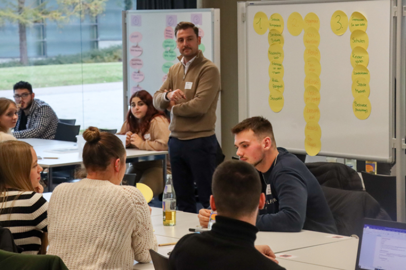 Eine Gruppe von Menschen sitzt an Tischen in einem hellen Seminarraum. Im Hintergrund sind Whiteboards mit bunten Klebepunkten und Listen zu sehe