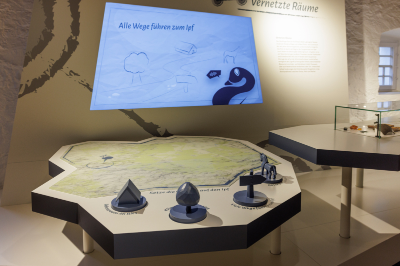 Ein Ausstellungsstück in einem Museum: Ein hexagonaler Tisch mit einer großen Karte oder Landschaftsmodell in hellen Tönen liegt flach auf der Tischplatte. Auf dem Tisch stehen mehrere kleine 3D-Modelle.