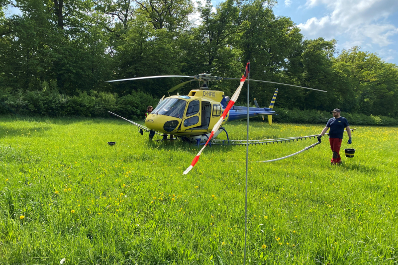 Grüne Wiese, darauf ein gelber Hubschrauber. Ein Mann breitet einen langen Schlauch aus.