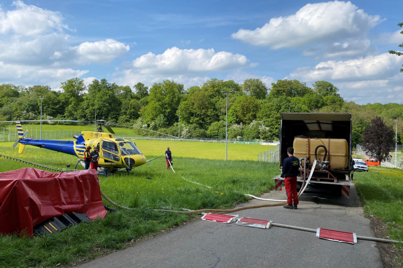 Größeres Gelände mit Geländewagen-Hubschrauber im Hintergrund. Ein gelbes Hubschrauberfahrzeug steht auf einer Wiese neben einem geschotterten Weg.