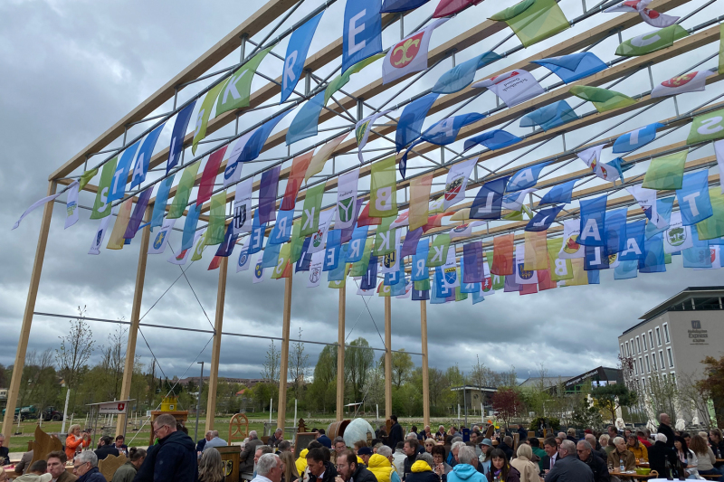 Außenaufnahme eines Kreispavillons, von der bunte Flaggenbänder in Blau, Grün, Gelb und Rot hängen.