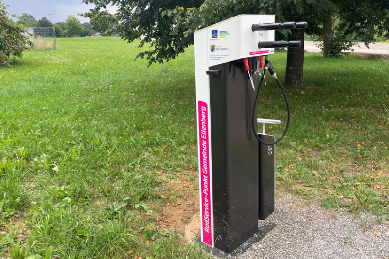 Wiese mit Baum, davor eine geschotterte Fläche mit einer Stele 'RadService-Punkt', an der Werkzeuge für die Fahrrad-Reparatur befestigt sind.