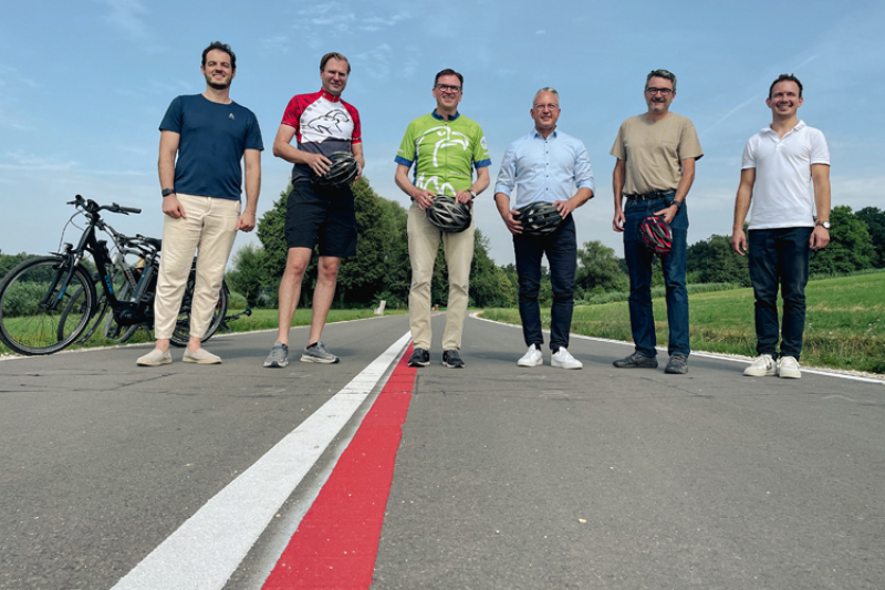 Auf einem Radweg mit einem rote und weißen Streifen stehen 6 Personen. Sie eröffnen eine neue Zählstelle für Radfahrende. In der Hand halten sie Fahrradhelme. Wiesen und Bäume im Hintergrund und zwei Fahrräder. 