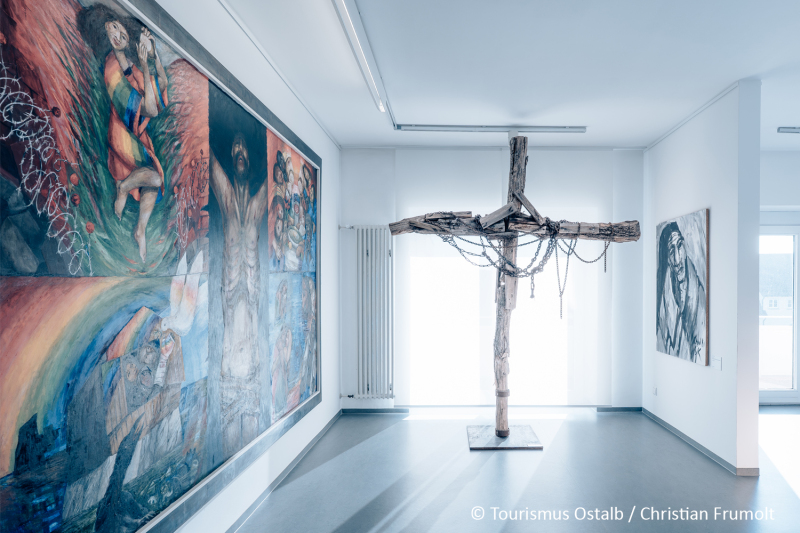 Großformatige Gemälde von Sieger Köder, vor einem Fenster steht ein großes Kreuz, umwickelt von einer Kette
